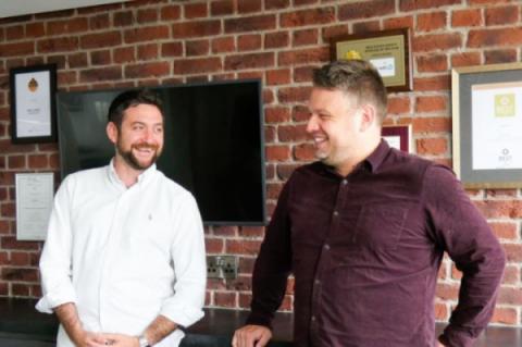 Two young men stand in a modern office, smiling. They both wear smart shirts.