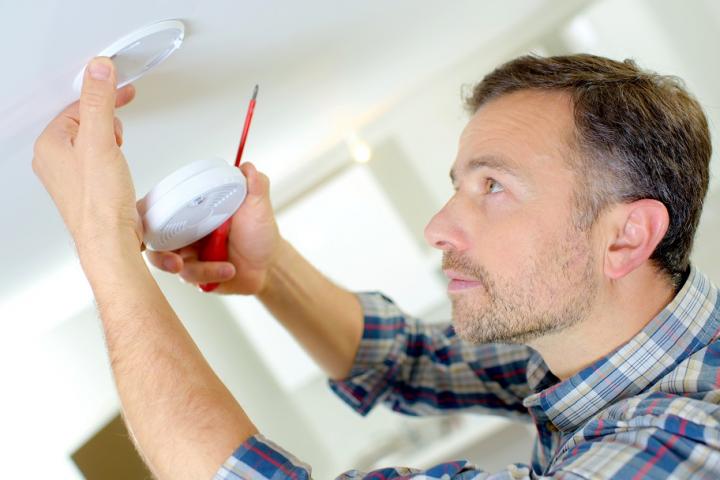 A man in a checky shirt looking up at smoke alarm with red screwdriver in hand