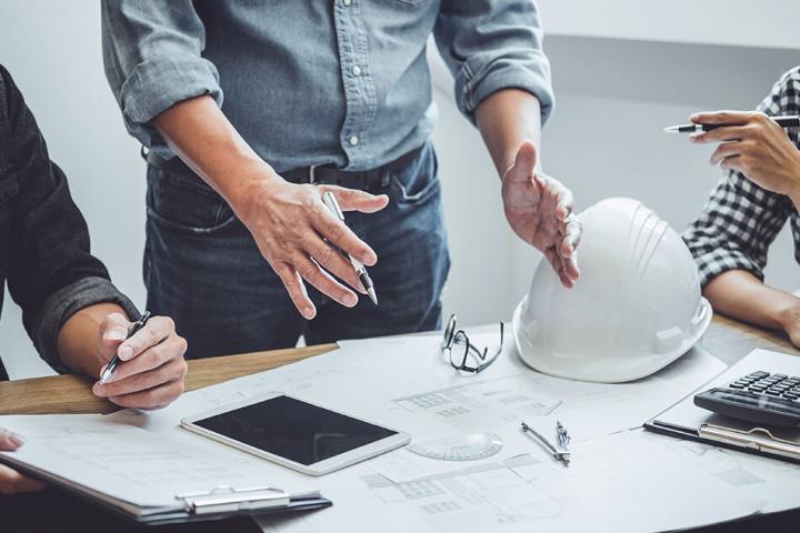 table with hard hat and papers on it. 3 workers stand over it