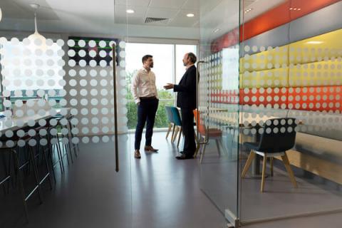 Camera shooting through a glass door at two men standing, having a conversation in a brightly coloured room
