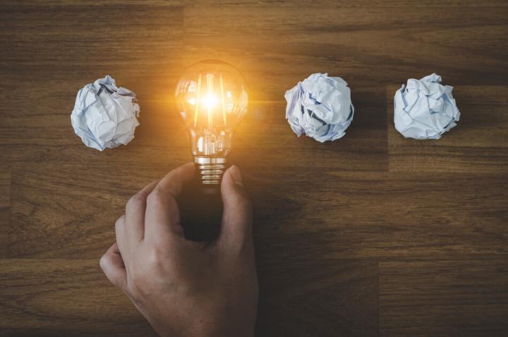 3 scrunched up pieces of paper flat on a wooden surface, in the middle replacing the 4th is a lightbulb