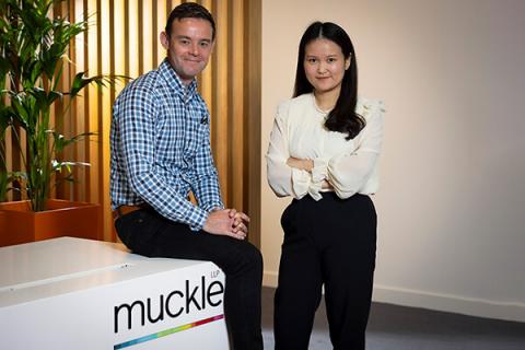 Adam sitting on a desk with the muckle logo on it, Zi is standing next to him