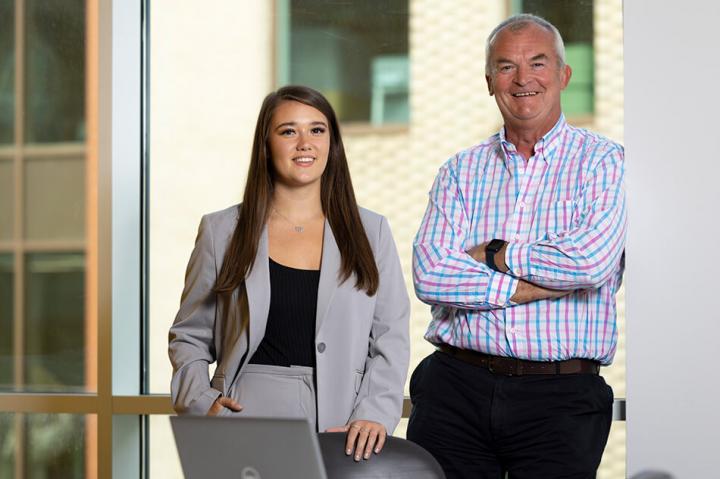 a woman and man standing left to right in front of a window with a laptop in the foreground 