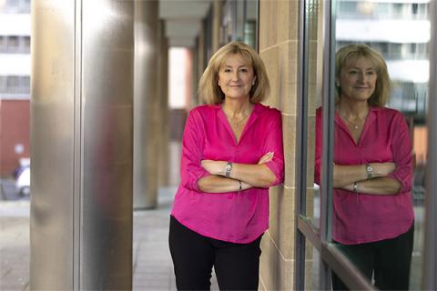 Susan Howe in a pink shirt leaning against a wall with a window reflection visible