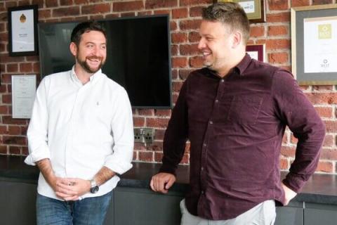 jamie cooke and ben ridgway standing next to each other with a brick wall in the background that has framed pictures on it