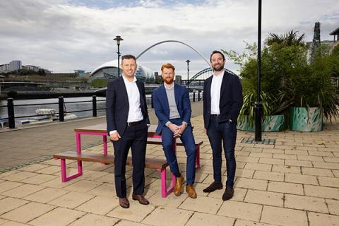Three men standing next to a bench on Newcastle's quayside with a bridge and river in the background