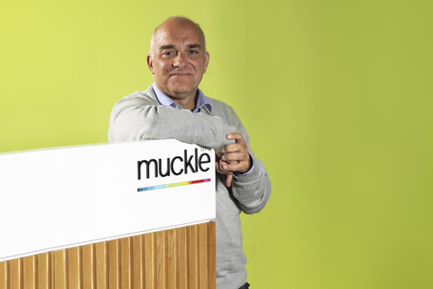 Jason Wainwright, managing partner leaning on a desk with a Muckle logo on it, green wall in the background