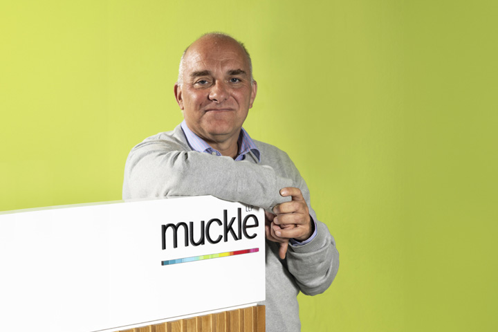 Jason Wainwright, managing partner leaning on a desk with a Muckle logo on it, green wall in the background