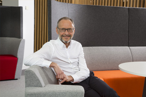 Hugh welch in a white shirt sitting on a orange and grey sofa, one arm over the back of the chair