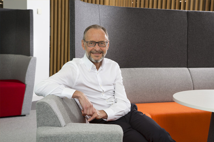 Hugh welch in a white shirt sitting on a orange and grey sofa, one arm over the back of the chair