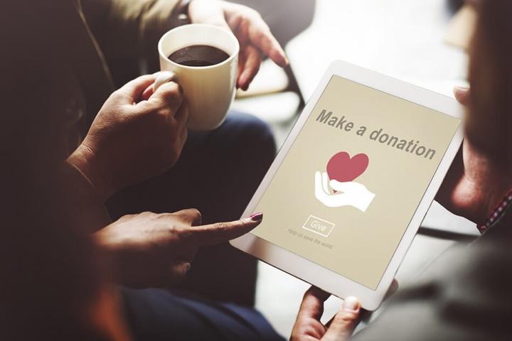 A person holding an ipad with the words make a donation on it and an illustration of a white hand holding a red heart. In the background there is another person holding a cup of coffee