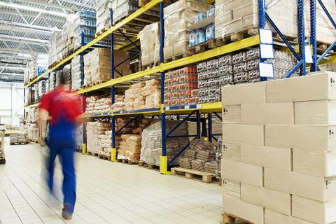 warehouse stacked with boxes on the shelf and a man wearing a red shirt and blue trousers walking away from the camera