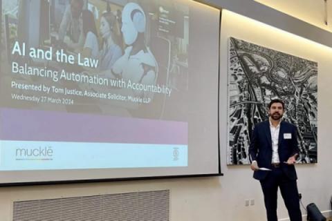 Tom justice, lawyer at Muckle LLP standing in front of a large projector screen with a presentation on it saying AI and the Law