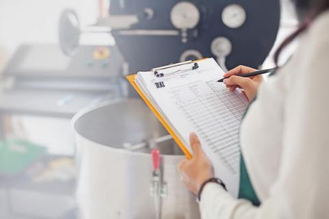 Woman in white coat holding a clipboard in front of machinery