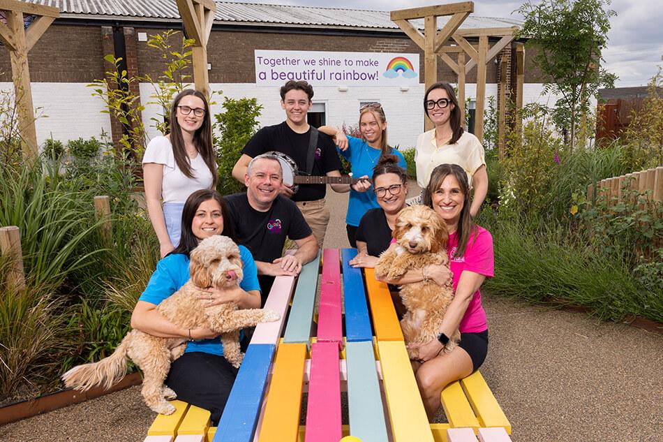 A group of 8 people and two dogs sitting around a colourful painted park bench