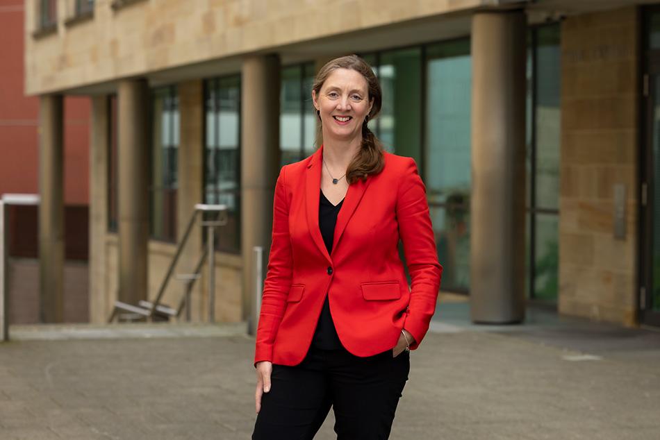 Alex Craig wearing a red jacket and black trousers standing in front of a building and smiling