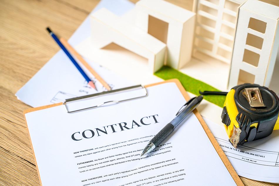 A construction contract on a table next to a tape measure and small-scale wooden buildings