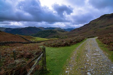 A valley in the Lake District with green grass and a stone path to the right, with mountains and a cloudy blue sky in the background