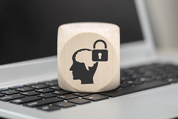 A wooden cube resting on a laptop keyboard. The cube displays the outline of a person's head with a padlock above it.
