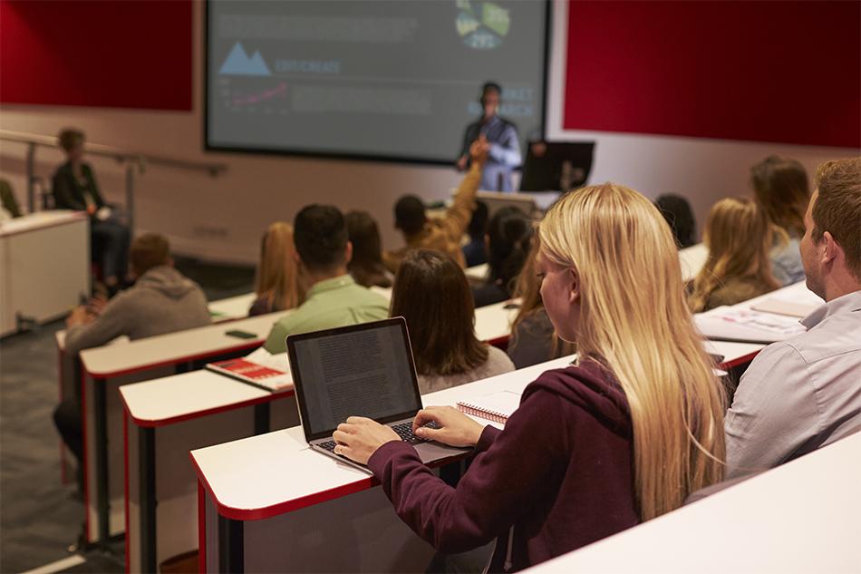 A group of students in a lecture theatre with red walls, with a lecturer visible in the background. In the foreground is a blonde female student working on a laptop.