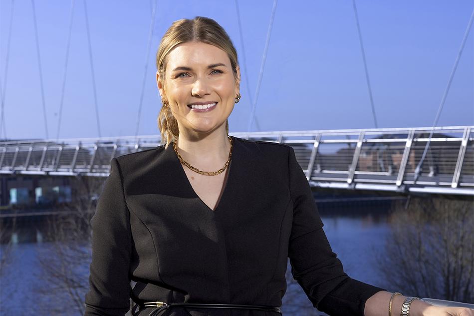 Headshot of Leah Duffield wearing a black jumpsuit standing in front of Teesside's Infinity Bridge