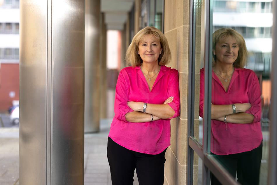susan howe wearing a pink shirt, leaning against a window, you can see her reflection in the window