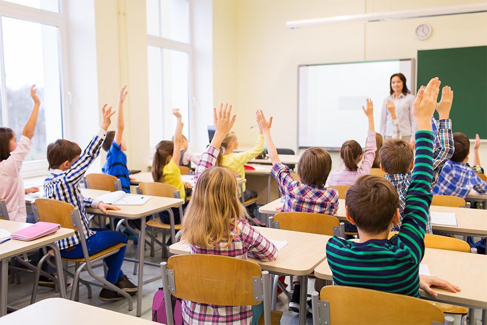 Schoolchildren in casual clothes sitting at desks with their hands raised, whilst a female teacher stands in front of them next to a whiteboard.
