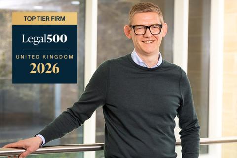 Close up on Ant Evans, in a dark grey jumper and blue shirt, leaning against a railing and smiling. The Legal 500 logo for top tier firm is superimposed to the left.