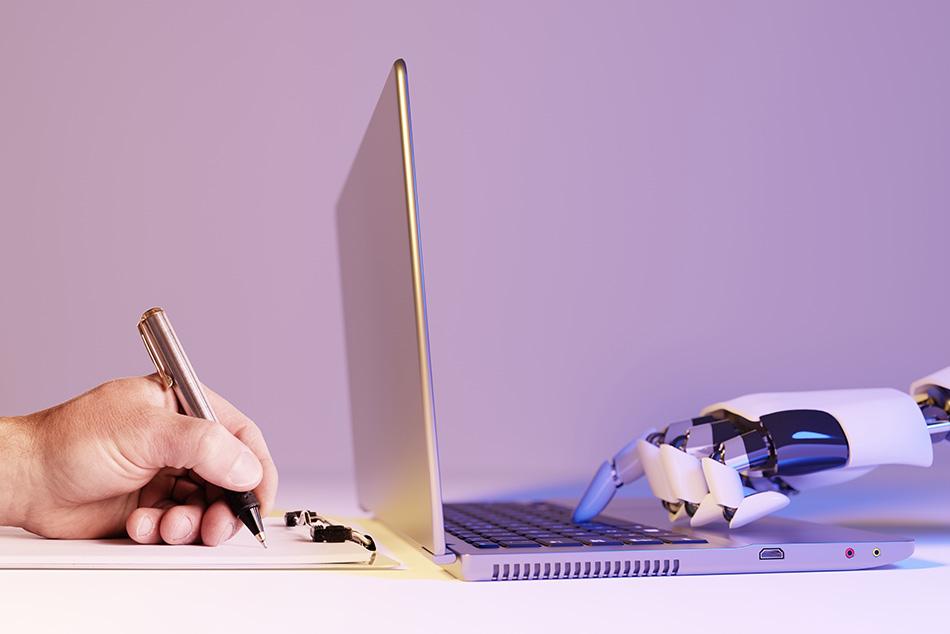 A robot hand typing on a laptop with a human hand writing on a notepad next to it