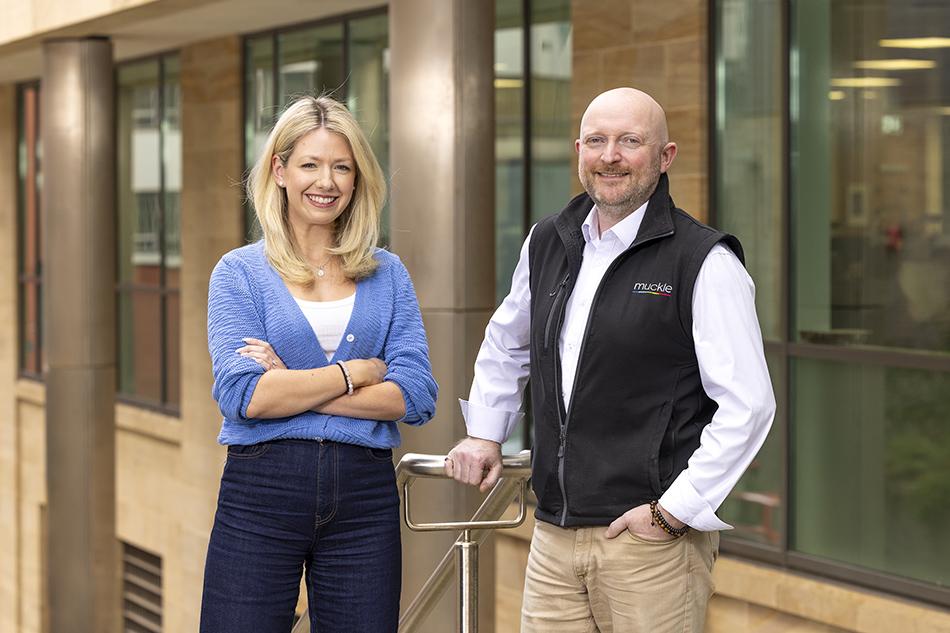 Kathryn Boyd, in a blue cardigan and blue jeans, and David Towns, in a white shirt, black gilet and neutral trousers, standing in front of a building and smiling