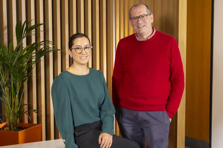 Phoebe Gogarty, wearing teal, and Chris Maddock, wearing dark orange, smiling at the camera with a plant in the background