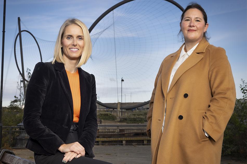 Rachel List, wearing an orange top and black jacket, and Gail Bennett, in a camel coat, standing in front of the Temenos monument in Teesside