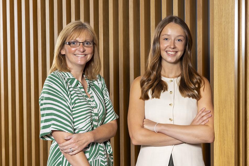 Joanne Davison, wearing a green and white stripy dress, and Jessie Melory, wearing a cream vest, standing in front of a panelled wall and smiling