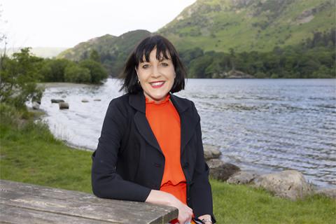 Rachael Stephenson, wearing a red dress and black jacket, sitting at a table in front of Ullswater Lake
