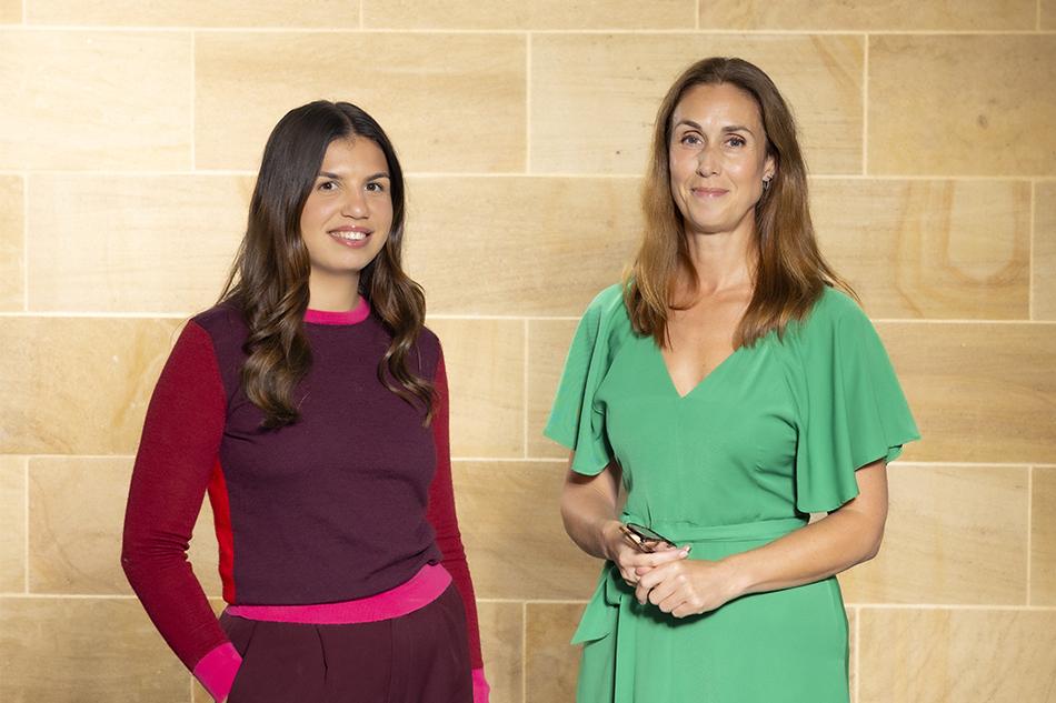 Ella Johnson, in a red and pink top, with Charlotte McMurchie, in a green dress, both standing in front of a beige tiled wall
