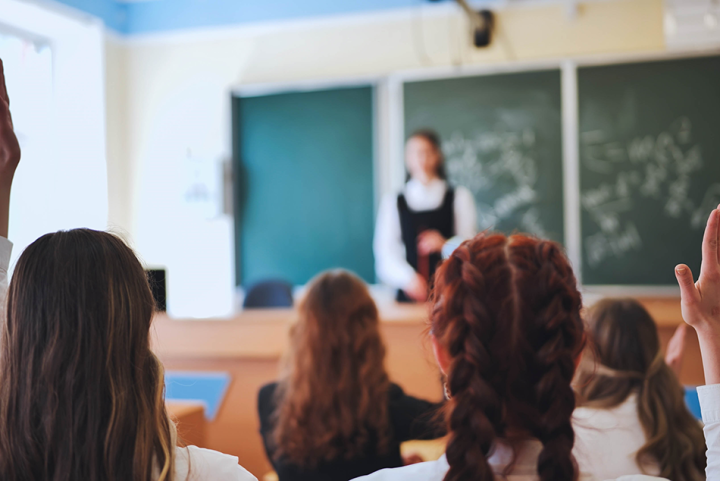 Close up of children in a classroom (backs to camera) with a teacher and blackboard visible in the background