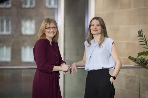 Joanne Davison, in a maroon dress, and Anna Glover, in a powder blue top and dark trousers, leaning on a railing and smiling at the camera