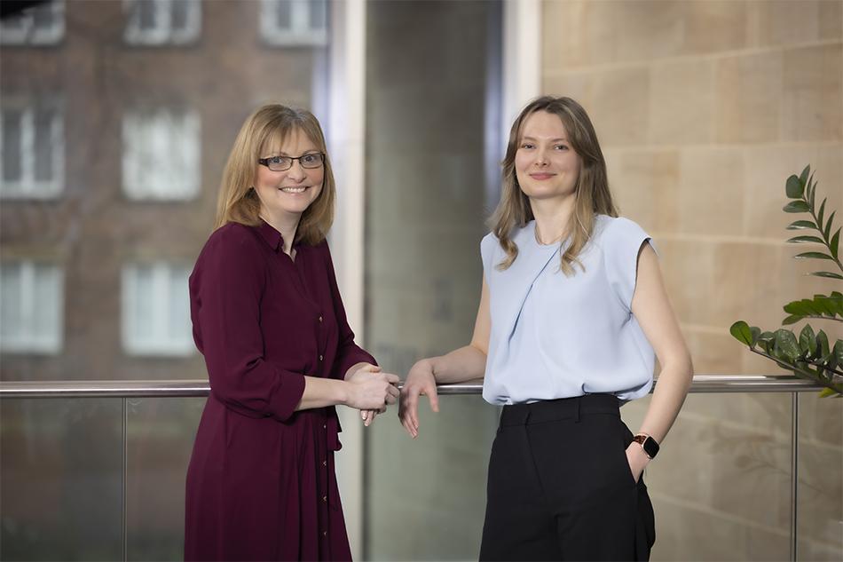 Joanne Davison, in a maroon dress, and Anna Glover, in a powder blue top and dark trousers, leaning on a railing and smiling at the camera