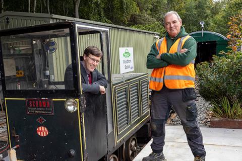 Man in suit sits in locomotive. Man in high-vis stands on the platform beside.