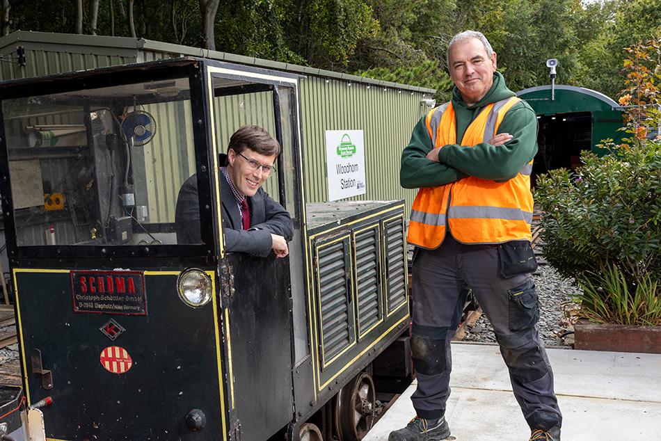 Man in suit sits in locomotive. Man in high-vis stands on the platform beside.